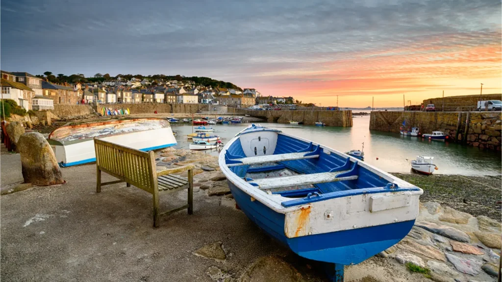 A picturesque harbor scene at sunset, featuring a row of upturned boats on the shore, a calm water surface dotted with boats, and a quaint village on the hill under a vibrant sky.