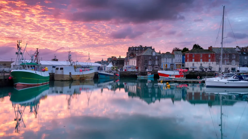 Sunset over a calm harbor featuring colorful clouds, reflective water, and moored boats with a backdrop of quaint buildings along the waterfront.
