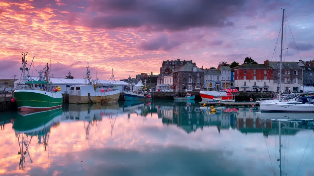 A tranquil harbor scene at sunset with vibrant sky reflections in calm water, featuring fishing boats, a sailboat, and colorful waterfront buildings.