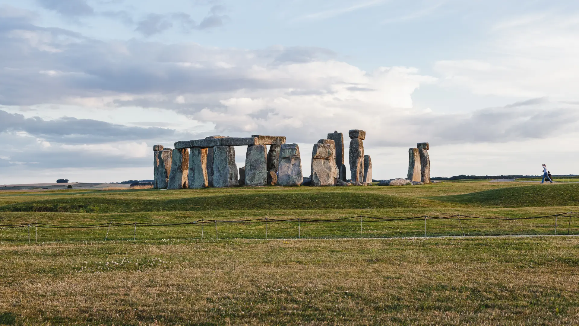 Ancient stonehenge monument with large standing stones arranged in a circle, under a vast sky with scattered clouds, surrounded by a grassy landscape. a lone visitor is visible in the distance.