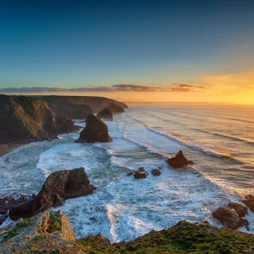 Sunset over a rugged coastline with cliffs and rock formations, waves crashing against the shore and reflecting the sunlight, under a clear sky.