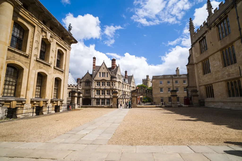 A sunny view of an ancient cobblestone square surrounded by historic academic buildings with distinctive architectural features, under a partly cloudy blue sky.