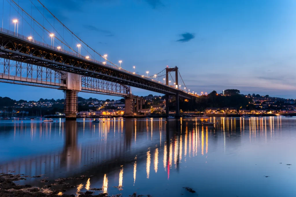 A serene twilight scene of a suspension bridge illuminated by lights with reflections on the calm water below, and a backdrop of a hillside village with glowing lights.