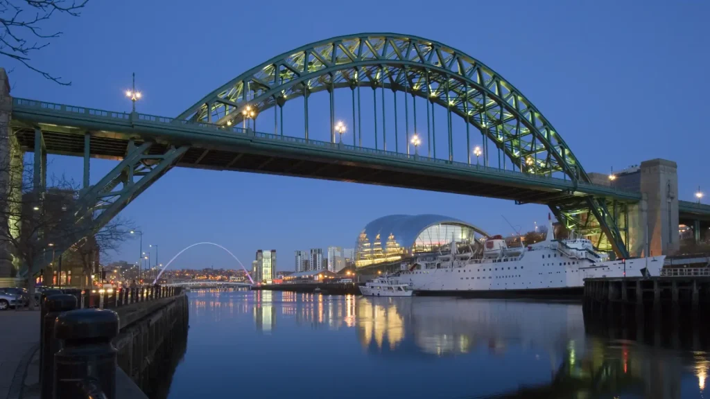 Evening view of the tyne bridge, a green arched bridge over the river tyne in newcastle, with illuminated sage gateshead building and lit cityscape reflected in the water.