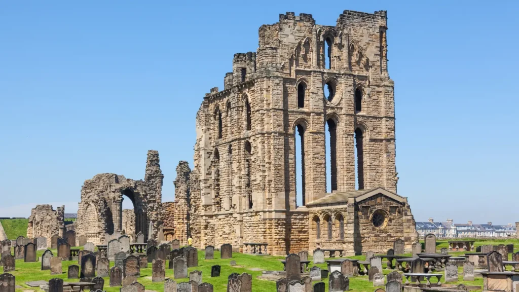 Ancient stone ruins of tynemouth priory and castle against a clear blue sky, surrounded by gravestones on green grass.