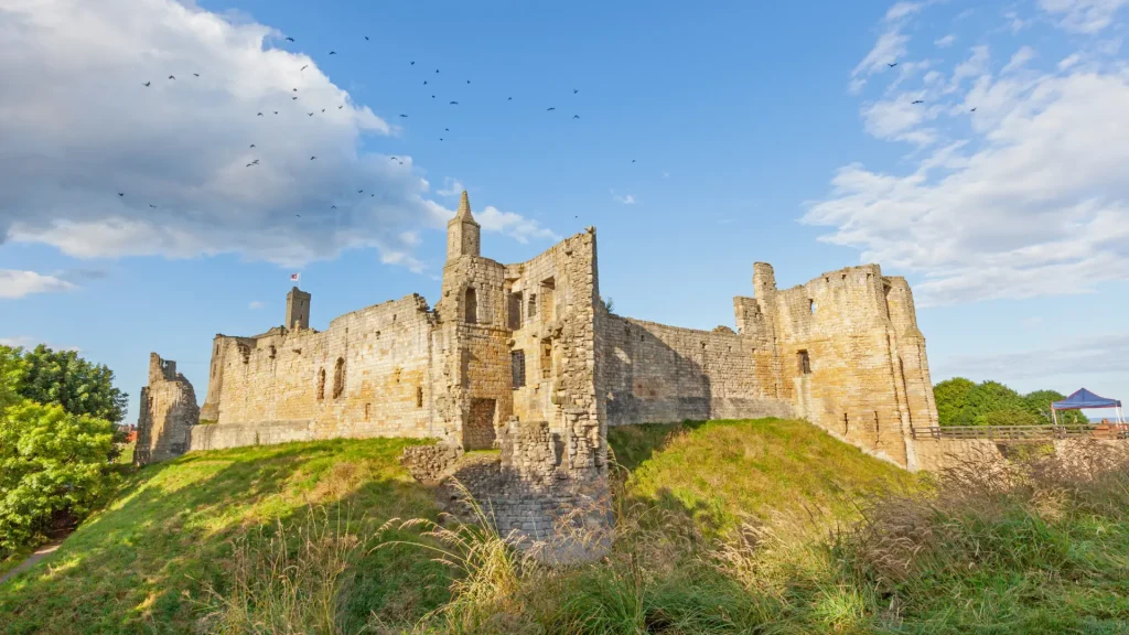 Ruins of a medieval stone castle on a grassy hill under a blue sky with scattered clouds and flying birds.