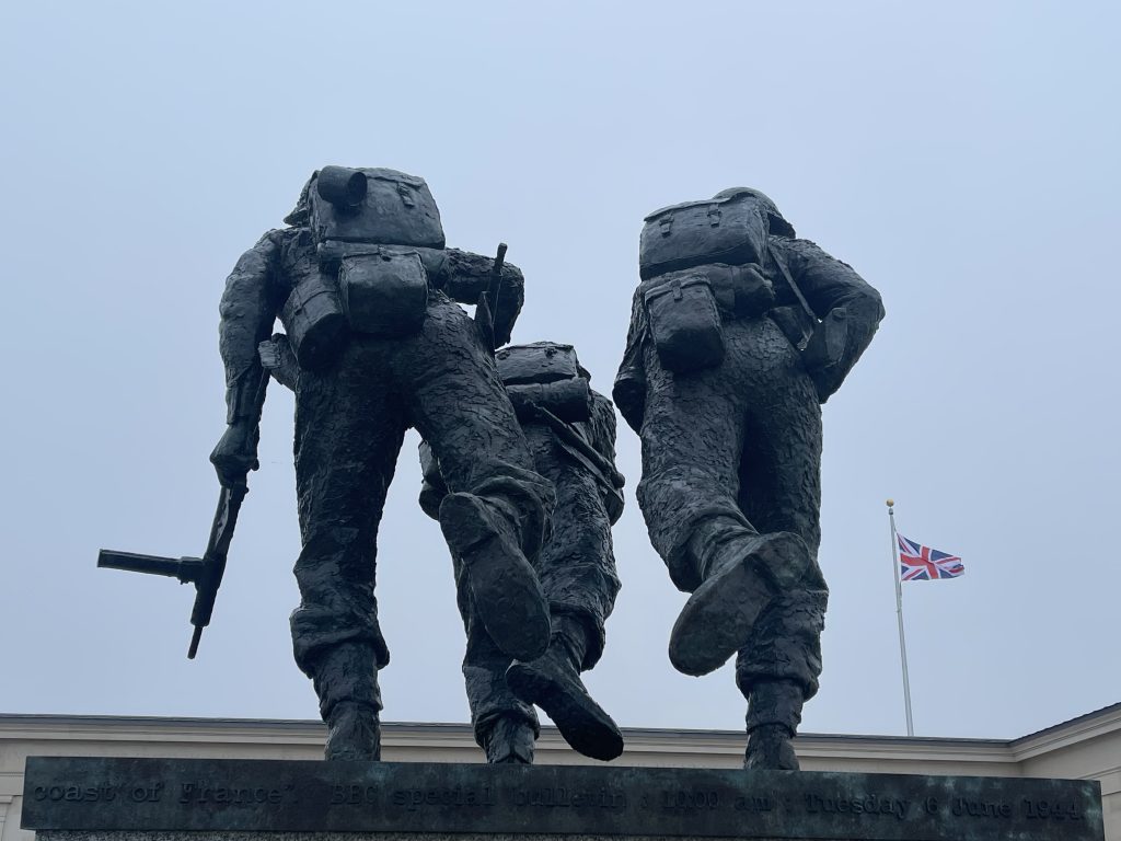 Bronze sculpture of three soldiers in uniform with large backpacks, monumentally depicted walking forward, set against a clear sky with a united kingdom flag fluttering in the background.