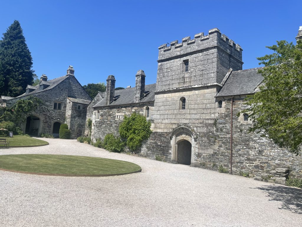 An ancient stone castle with a prominent gatehouse in the center, surrounded by lush greenery under a clear blue sky. a gravel path leads towards the gate.