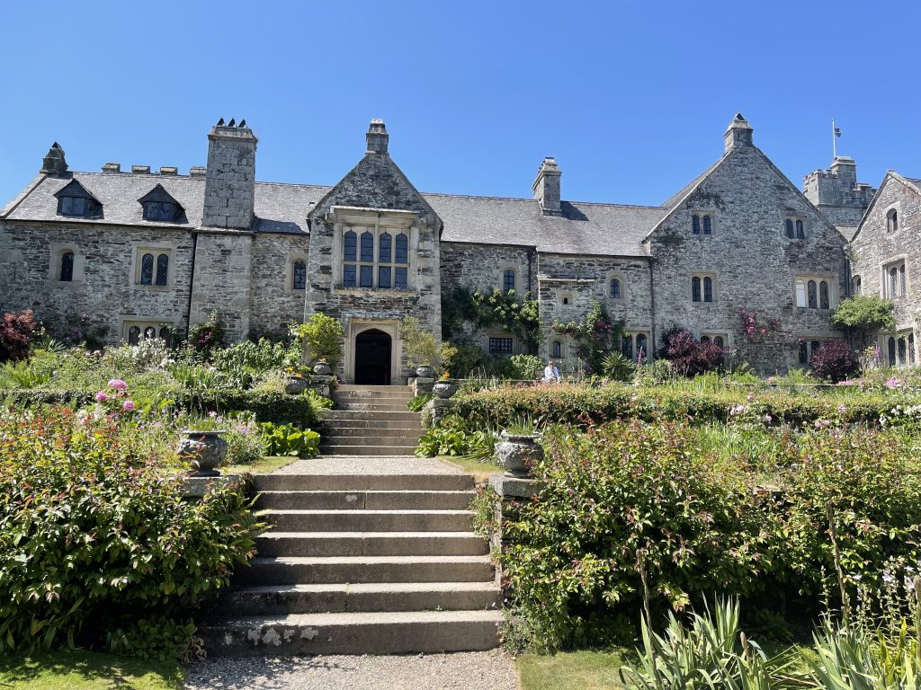 A grand stone building with gothic-style windows surrounded by lush gardens under a clear blue sky. stone steps lead up to the main entrance, flanked by vibrant flowers and greenery.