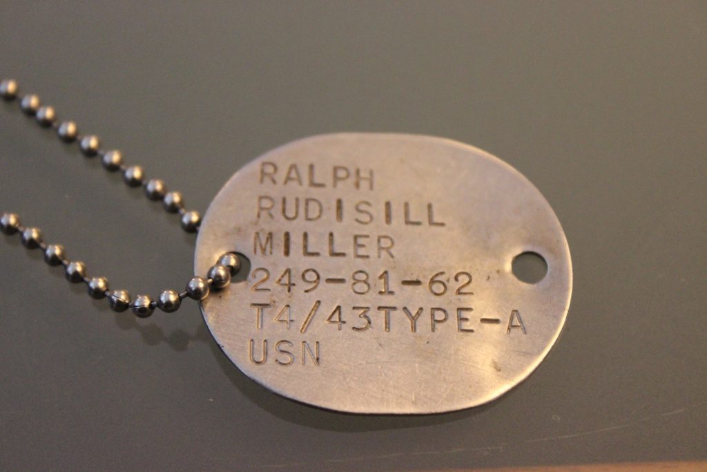 A military dog tag on a gray surface with engraved text that includes the name "ralph rudisill miller," a serial number, blood type, and "usn" indicating u.s. navy affiliation.