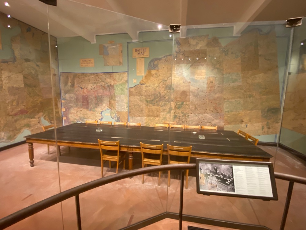 A historical display featuring a large, long table surrounded by chairs, set in front of a wall-sized, faded map titled "battle map." exhibit encased in glass with informative plaques visible.