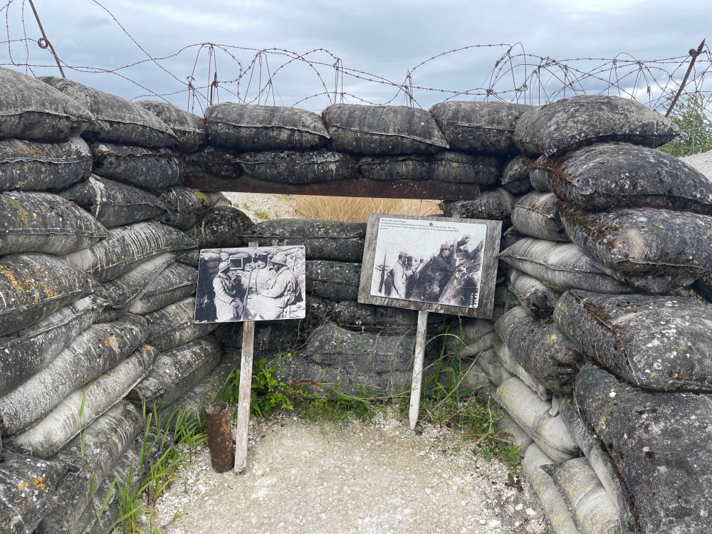 Historical photos mounted on boards beside a sandbag and barbed wire barrier under a cloudy sky, evoking a wartime scene.