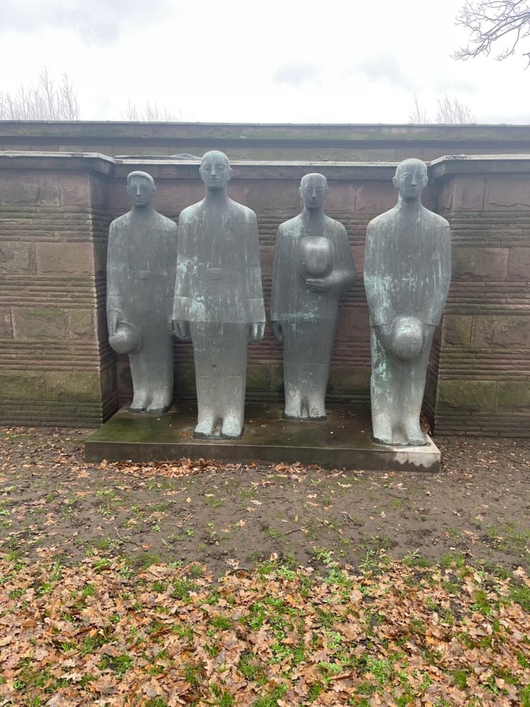 Four stone statues standing in a row against a wooden wall, each figure with a unique, stylized pose. the setting includes a ground covered with autumn leaves.
