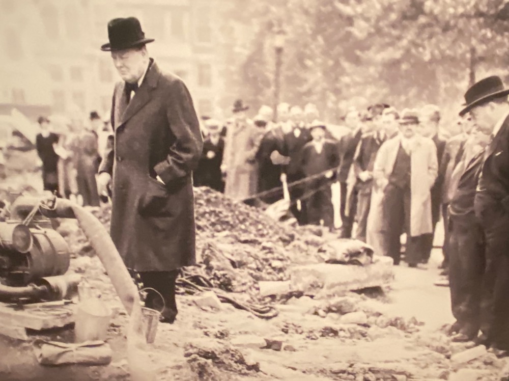 A black-and-white historical photo depicting a man in a top hat inspecting a construction site, with observers lined up behind barriers in the background.