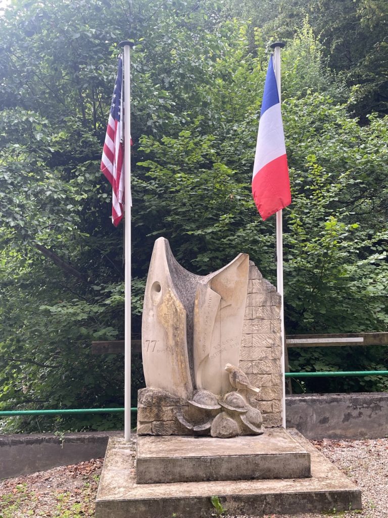 A stone memorial sculpture with a large eagle and flowers, flanked by an american flag on the left and a french flag on the right, located in a lush forest setting.