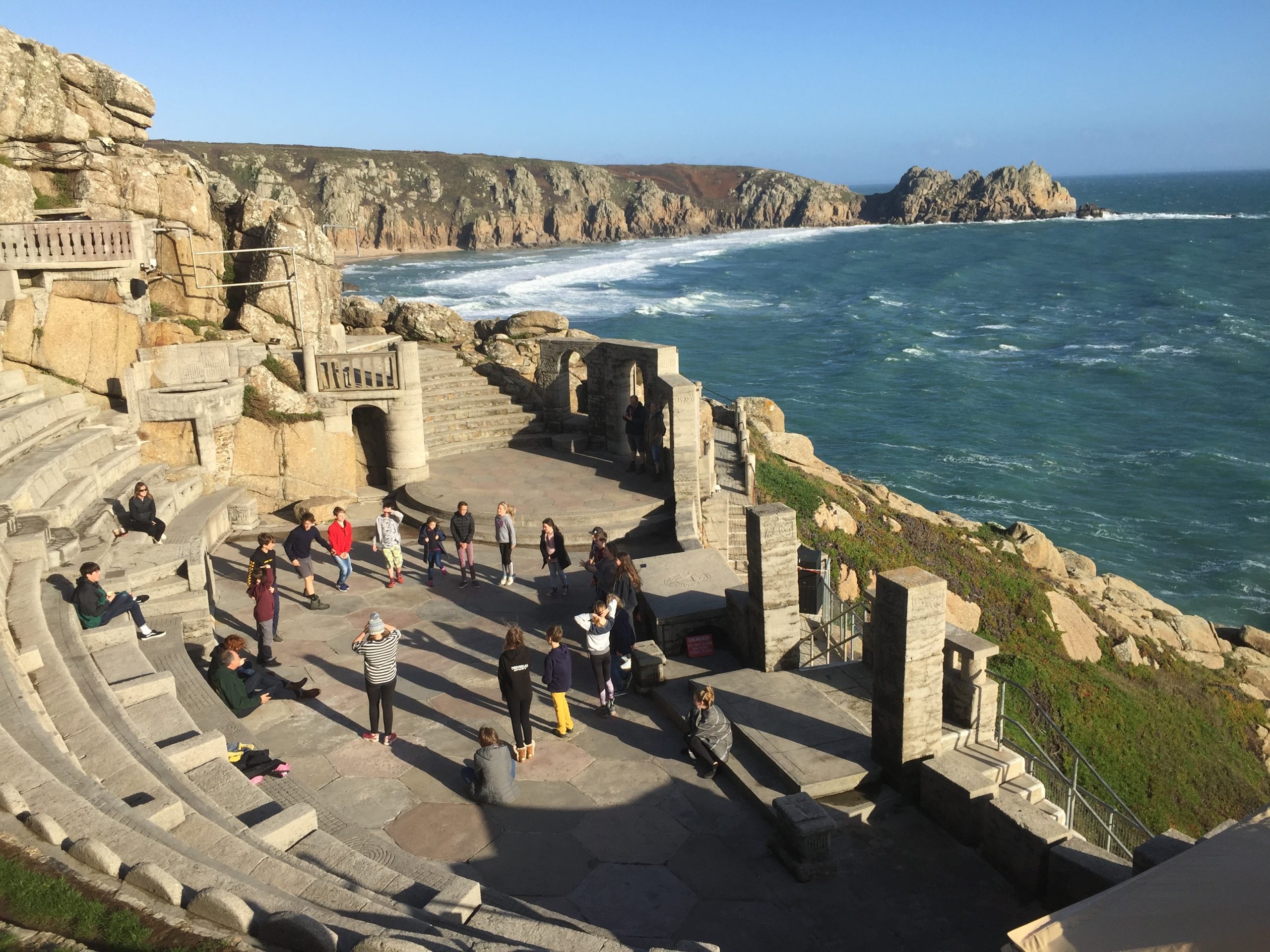 A coastal amphitheater with people sitting on stone benches and others standing, overlooking a choppy sea surrounded by rugged cliffs under a bright blue sky.