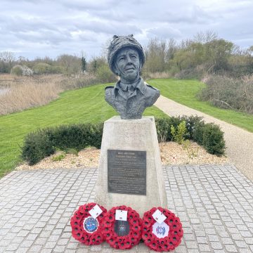 A bronze bust of a woman wearing a headscarf, displayed on a pedestal with a plaque and flanked by three wreaths with poppies. the monument overlooks a park path and a serene pond.