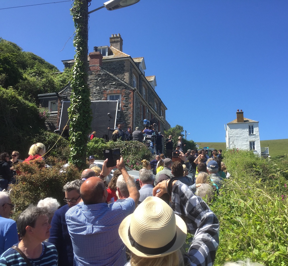A crowd of people outdoors, many taking photos, with a focus on a stone house nestled amongst lush greenery under a clear blue sky.