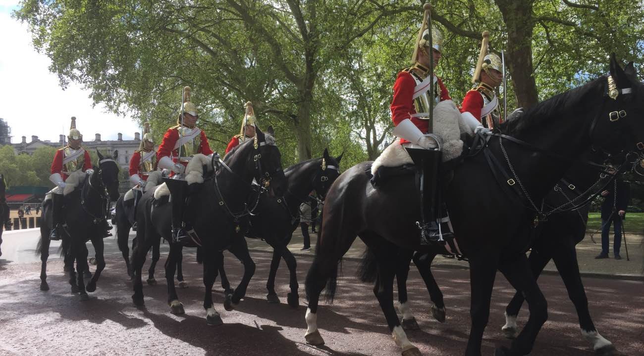 A group of ceremonial horse guards dressed in red and white uniforms riding black horses in a park-like setting, likely during a formal military parade.