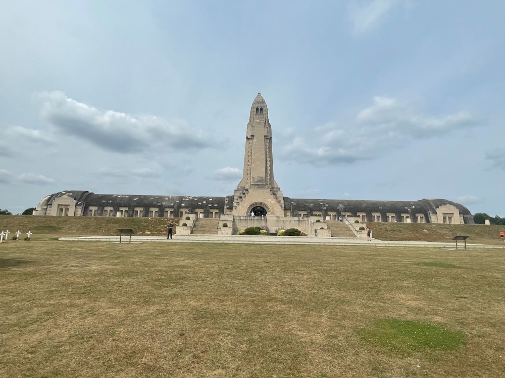 A wide-angle view of the douaumont ossuary in verdun, france, under a cloudy sky. the memorial, with its high tower and arched stone structure, stands in a large grassy area with a few visitors.
