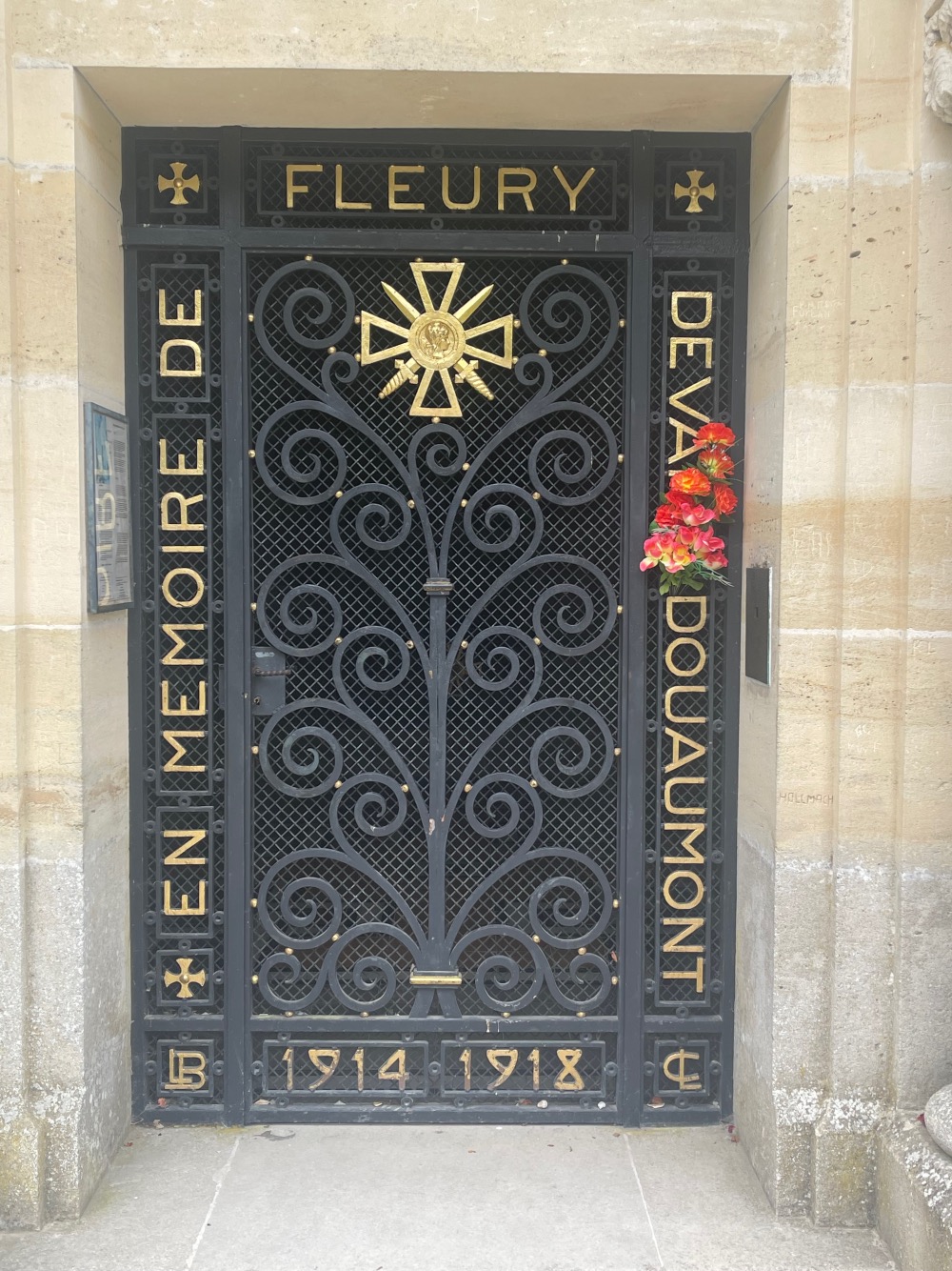 Black iron gate with intricate designs, featuring a gold star emblem at the top and inscribed with "fleury devant douaumont 1914 - 1918 en memoire de" above red flowers to the side.