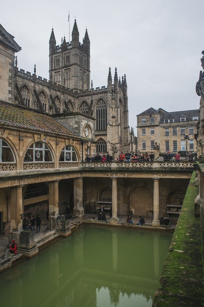 View of the historic roman baths in bath, england, featuring the green bath water, surrounding ancient stone architecture, and the bath abbey in the background.