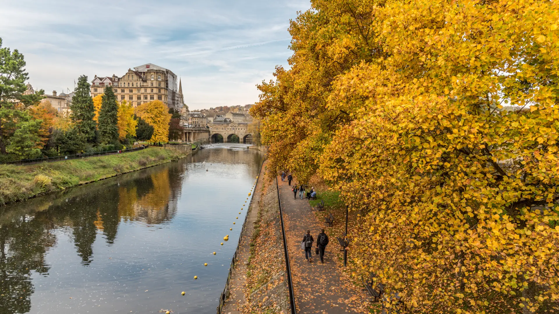 Autumn scene showing people walking along a tree-lined river path, with vibrant yellow foliage, overlooking a historic stone bridge and grand buildings in the background.