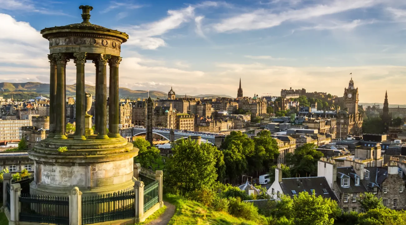 A panoramic view of edinburgh from calton hill at sunset, featuring the dugald stewart monument in the foreground and the city skyline with historic buildings.