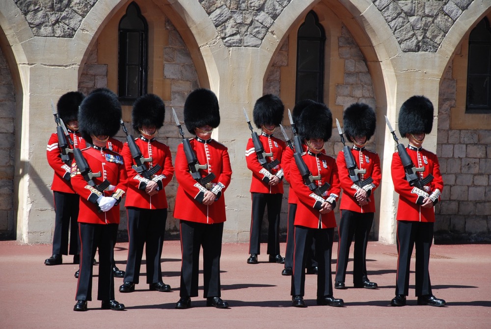 A row of eight guards in traditional red uniforms and tall black fur hats standing solemnly at a ceremony, with a historical stone building in the background.