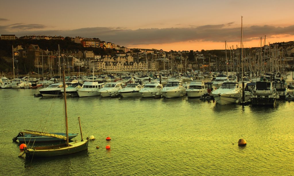A serene harbor filled with boats at sunset, water reflecting gold hues, with a hilly town in the background under a dusky sky.