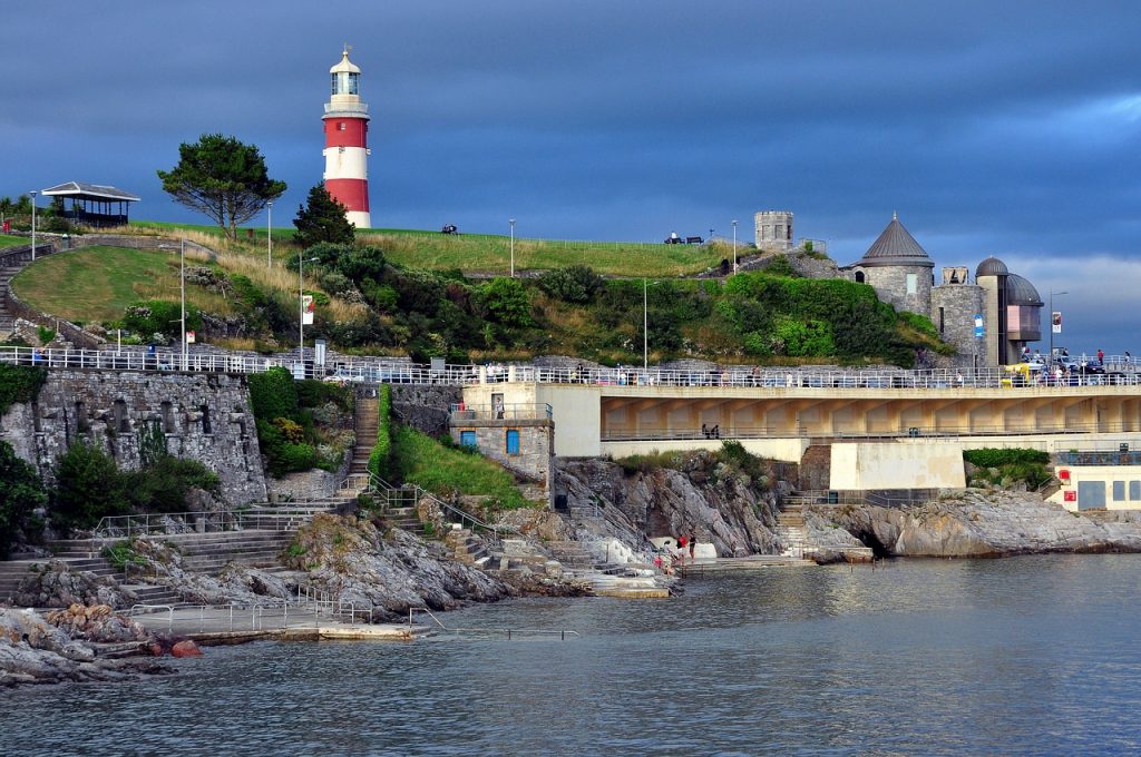 A scenic coastline with a red and white lighthouse atop a green hill, featuring a stone bridge, historical buildings, and rocky shore under a cloudy sky.