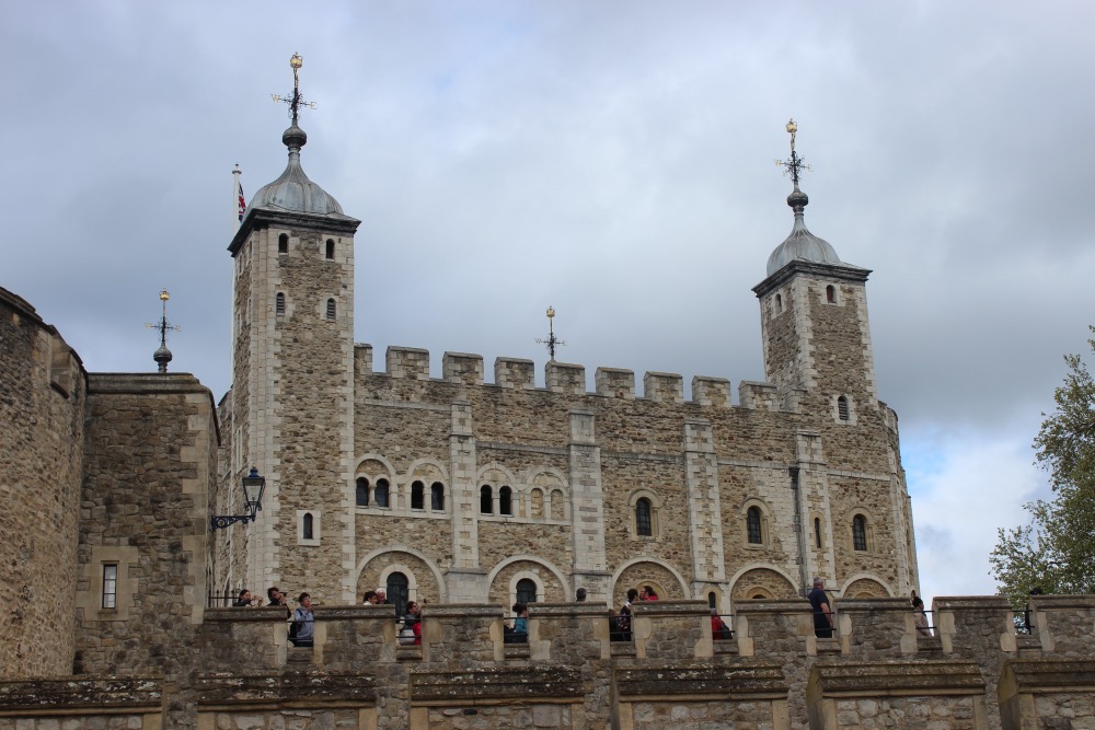 The image shows the historic exterior of the tower of london, featuring two prominent towers with weather vanes and several arched windows. visitors are seen on a balcony admiring the view.