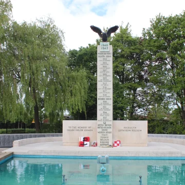 A war memorial with a vertical column topped by an eagle, flanked by weeping willow trees and fronted by a reflective pool, against a cloudy sky.
