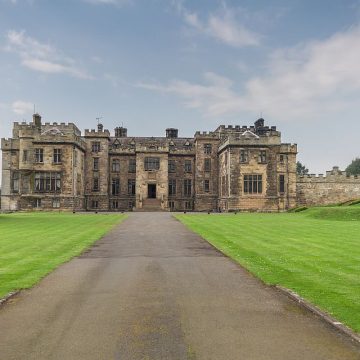 A historic stone castle with lush green lawns, featuring a central path leading to the ornately detailed facade, under a cloudy sky.