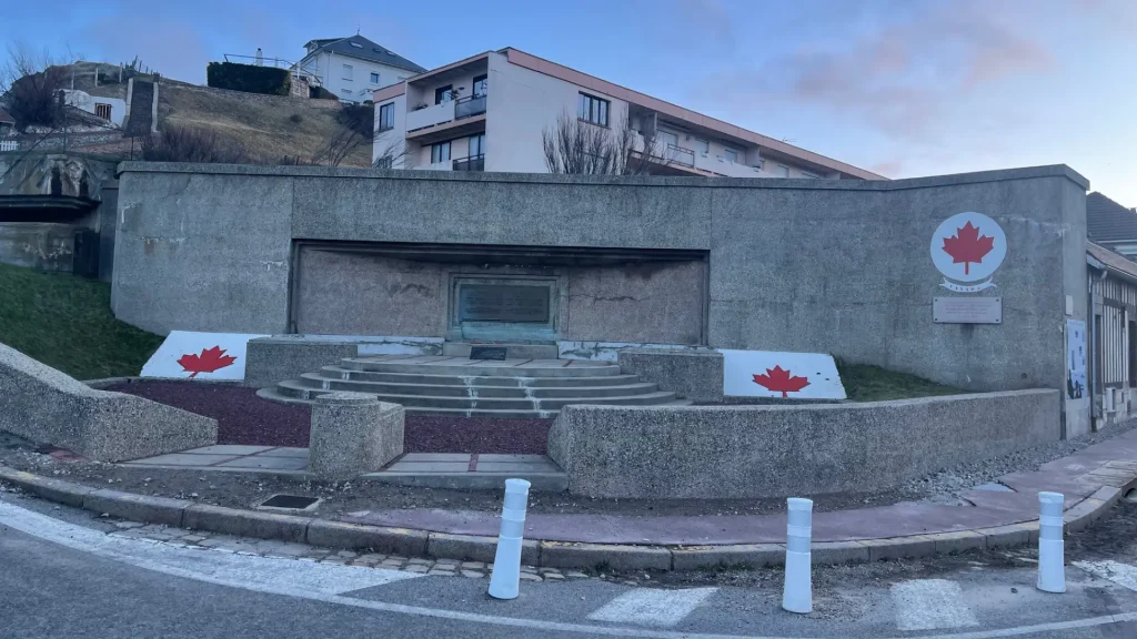 A memorial site with flag emblems and steps leading up to a central bronze plaque, located on a curved street corner with buildings in the background under a clear blue sky.