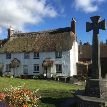 A charming, white, thatched-roof cottage with bright flowers in the foreground and a stone cross on the right, set against a backdrop of other houses under a clear blue sky.