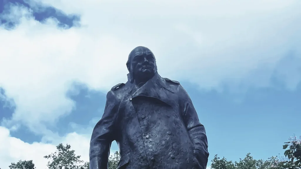 A statue of a stern-looking man with a robust build, wearing a military-style coat under a cloudy sky.