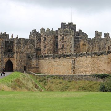 A grand medieval castle with multiple battlements and a large arched entrance. people are visible walking towards it on a grassy path, with a cloudy sky overhead.