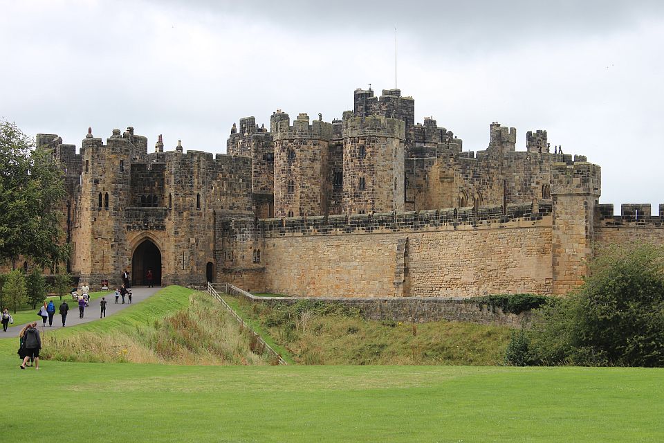 A grand medieval castle with multiple battlements and a large arched entrance. people are visible walking towards it on a grassy path, with a cloudy sky overhead.