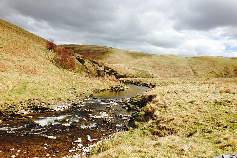 A scenic view of a meandering stream flowing through a valley with grassy hills under a partly cloudy sky. trees line the stream and a stone wall runs alongside.