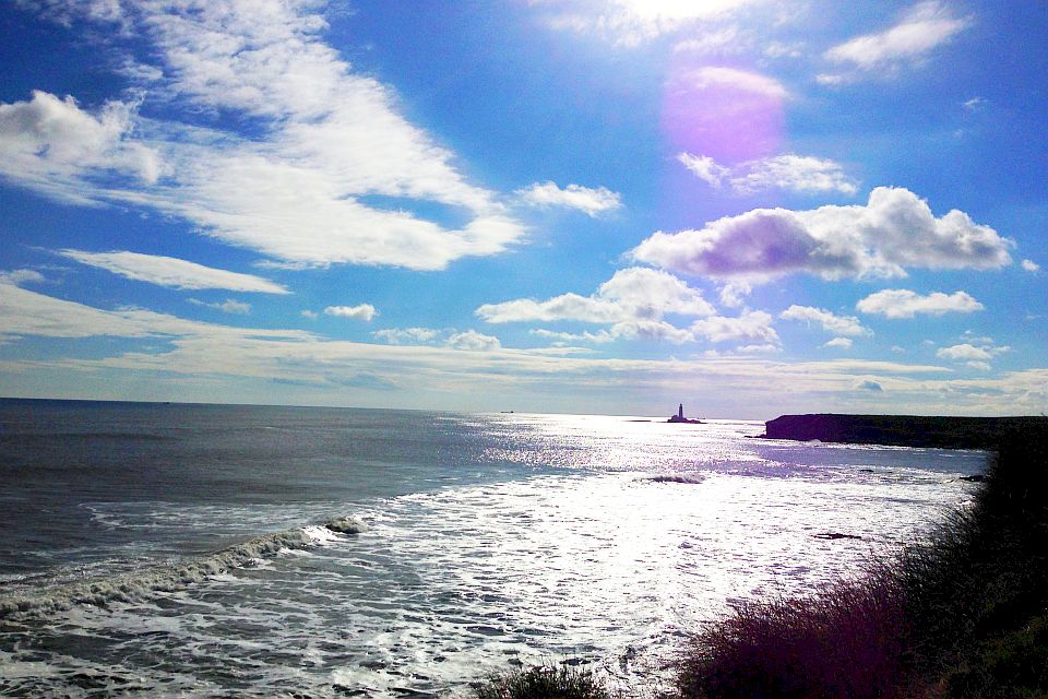 A scenic view of the ocean with waves hitting the shore under a blue sky dotted with clouds and sunlight reflecting off the water's surface. a distant lighthouse is visible on the horizon.