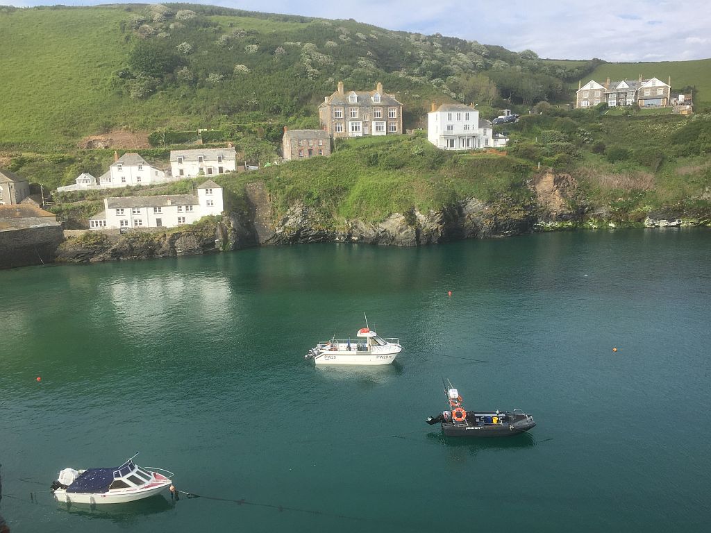 A scenic coastal view featuring a calm bay with three boats—two with white hulls and one black—in the foreground. lush green hills and a row of traditional houses overlook the water.