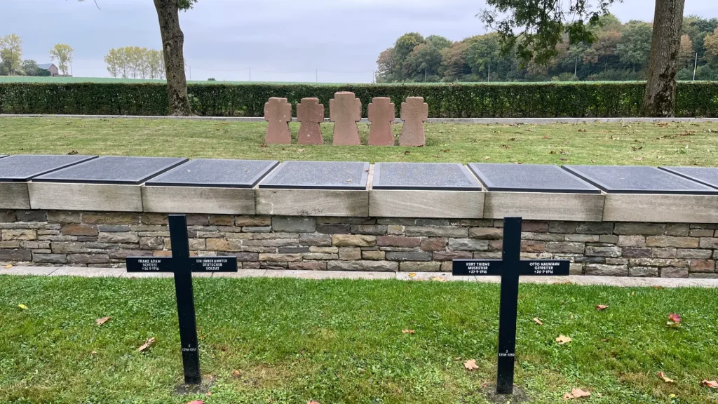 A somber memorial with black crosses, bronze plaques, and stone tablets displaying names, set against a serene park backdrop with lush greenery and a gray sky.