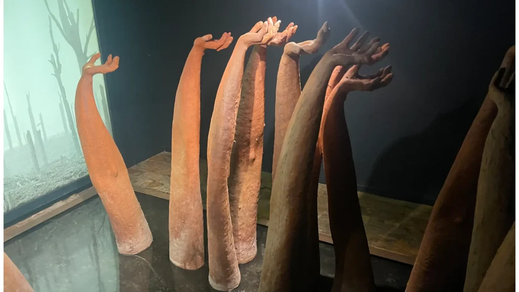 Sculptural installation of various elongated, vertical forms resembling organic tree branches or limbs, with hand-like extensions at the top, presented in a dimly lit gallery setting.