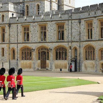 Guards in traditional red and black uniforms march in front of a historic castle with ornate stonework and large windows. one guard stands alone, watching.