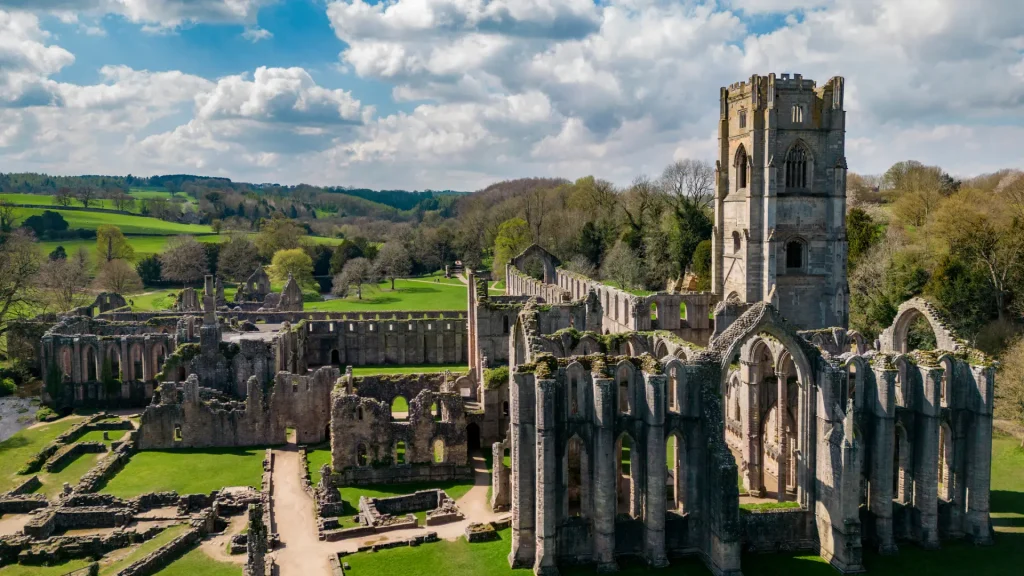 Aerial view of the ruins of a grand medieval abbey with an intact tower, surrounded by lush green fields under a partly cloudy sky.