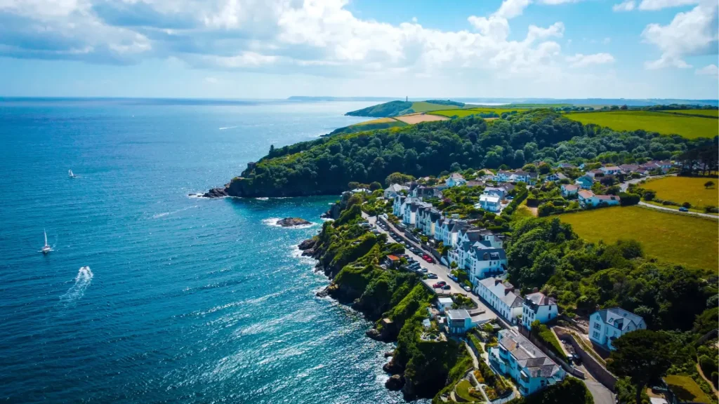 Aerial view of a coastal village with white houses lined along a curving road beside the sea, surrounded by green hills and a clear blue sky.
