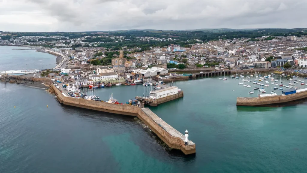 Aerial view of a coastal town with curved breakwaters encircling a harbor filled with boats, adjacent to a densely populated town and rolling hills in the background.