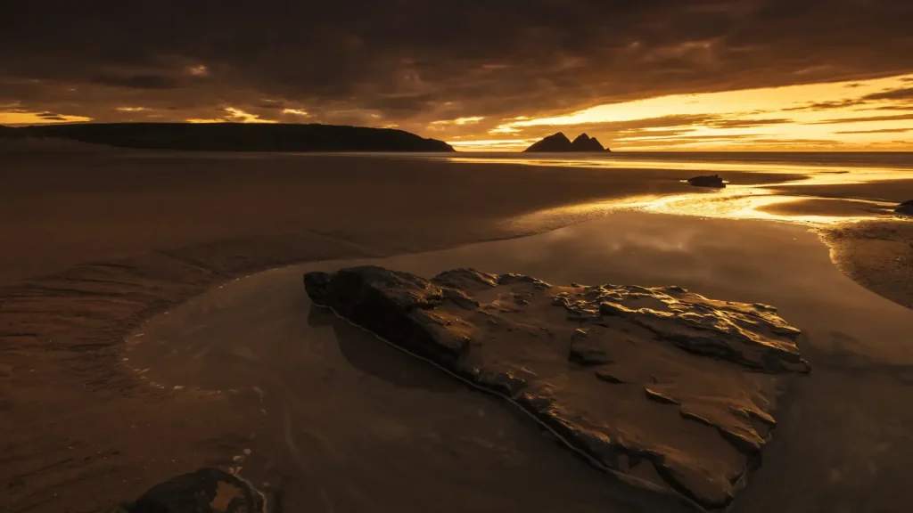 A serene sunset at a beach, showing vivid orange skies reflected in tidal water with prominent rocks in the foreground and distant islands on the horizon.
