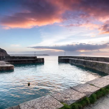 A serene harbor at sunset, featuring a calm sea bordered by stone walls, a small building to the left, and a vividly colored sky with clouds reflecting pink and purple hues.